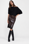 A model wearing a black off-the-shoulder knit top, a shiny dark brown faux-leather midi skirt, and black knee-high boots, posed against a light gray studio background.