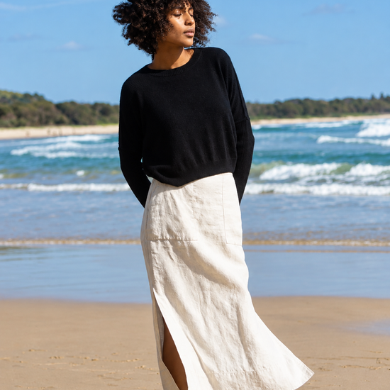 A woman stands barefoot on a sandy beach wearing a black long-sleeve knit top and a flowing off-white midi skirt with a side slit, with ocean waves and blue sky in the background.
