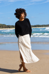 A woman stands barefoot on a sandy beach wearing a black long-sleeve knit top and a flowing off-white midi skirt with a side slit, with ocean waves and blue sky in the background.