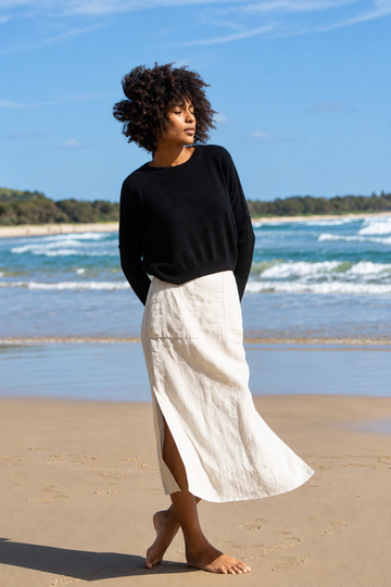 A woman stands barefoot on a sandy beach wearing a black long-sleeve knit top and a flowing off-white midi skirt with a side slit, with ocean waves and blue sky in the background.