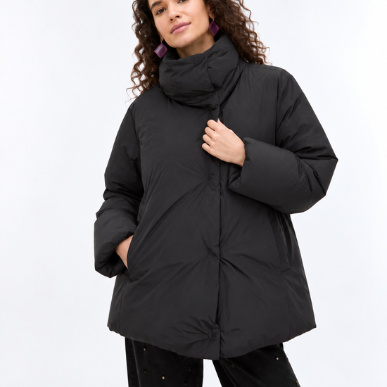 A woman wearing a black oversized puffer jacket with a high collar and front zipper, paired with black pants, photographed against a plain light gray studio background.