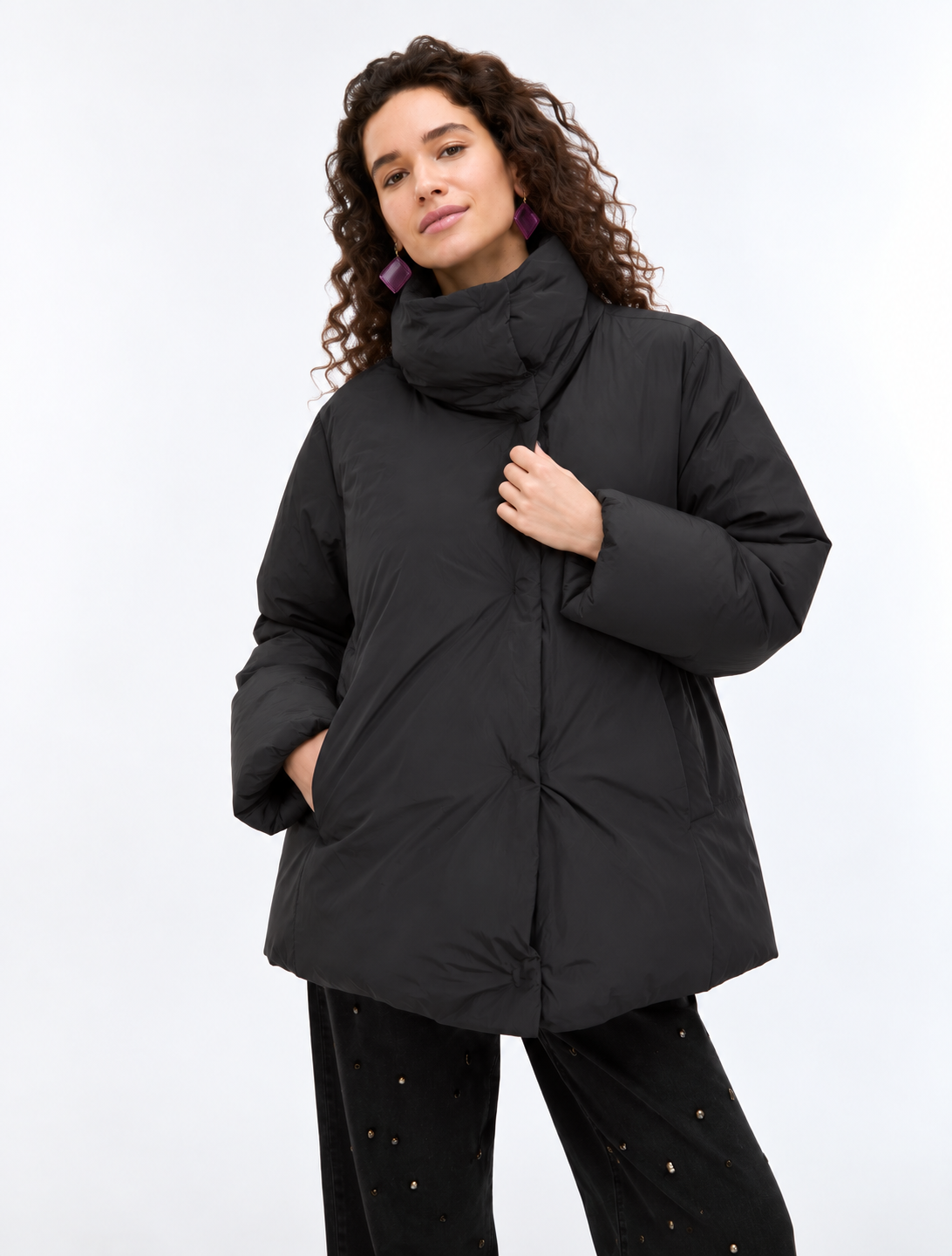 A woman wearing a black oversized puffer jacket with a high collar and front zipper, paired with black pants, photographed against a plain light gray studio background.