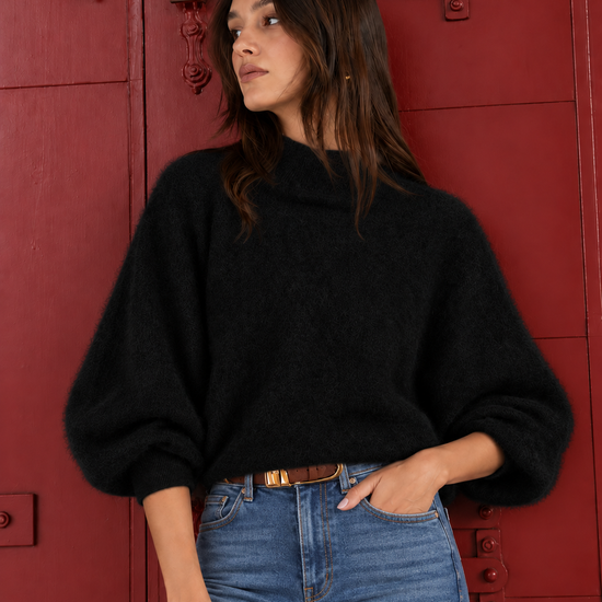 A woman wearing a black fuzzy knit sweater with balloon sleeves and blue jeans, standing in front of a red wooden door.