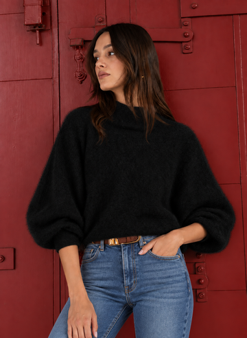 A woman wearing a black fuzzy knit sweater with balloon sleeves and blue jeans, standing in front of a red wooden door.