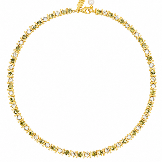 A gold tennis necklace featuring small green gemstones and clear stones, displayed on a clean white background. A gold tag is attached near the clasp.