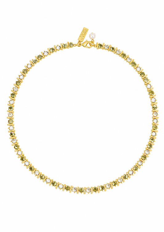 A gold tennis necklace featuring small green gemstones and clear stones, displayed on a clean white background. A gold tag is attached near the clasp.