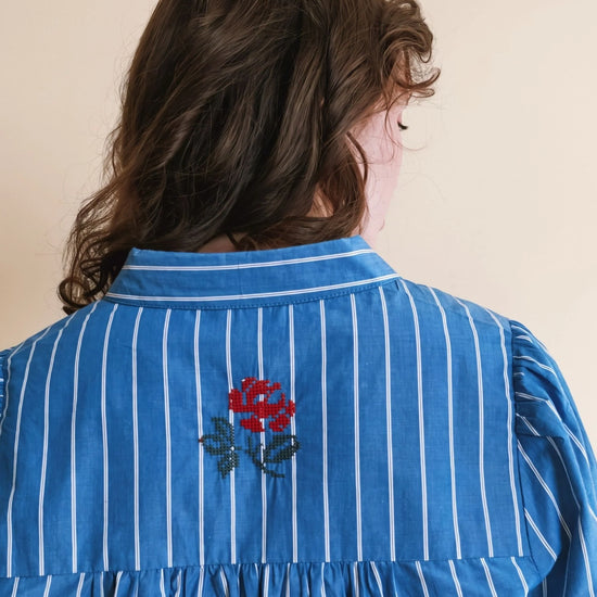 A person shown from the back wearing a blue long-sleeve blouse with thin white vertical stripes and a red floral embroidery centered on the upper back.