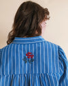 A person shown from the back wearing a blue long-sleeve blouse with thin white vertical stripes and a red floral embroidery centered on the upper back.