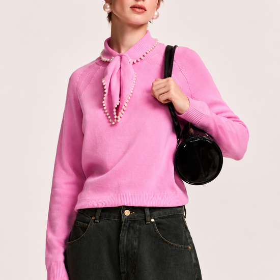 A model wearing a bright pink knit sweater with a pearl-trimmed tie-neck collar, paired with dark wide-leg jeans, a black baseball cap, and a black shoulder bag, set against a plain light background.