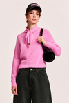 A model wearing a bright pink knit sweater with a pearl-trimmed tie-neck collar, paired with dark wide-leg jeans, a black baseball cap, and a black shoulder bag, set against a plain light background.