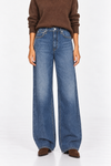 A model wearing a pair of wide-leg blue denim jeans, styled with a brown knit sweater and brown suede shoes, photographed against a clean white studio background.