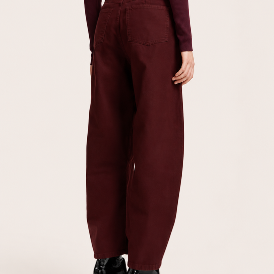 A model shown from the back wearing wide-leg maroon trousers, a dark burgundy top, a bright pink belt, and black pointed-toe heeled boots, set against a plain light beige studio background.