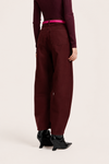 A model shown from the back wearing wide-leg maroon trousers, a dark burgundy top, a bright pink belt, and black pointed-toe heeled boots, set against a plain light beige studio background.