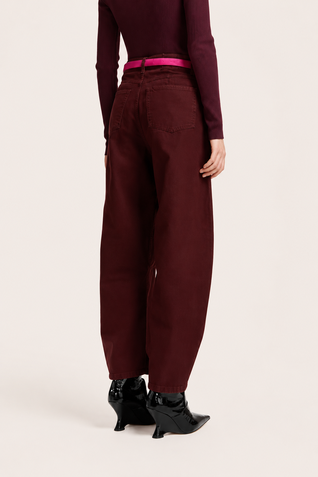 A model shown from the back wearing wide-leg maroon trousers, a dark burgundy top, a bright pink belt, and black pointed-toe heeled boots, set against a plain light beige studio background.