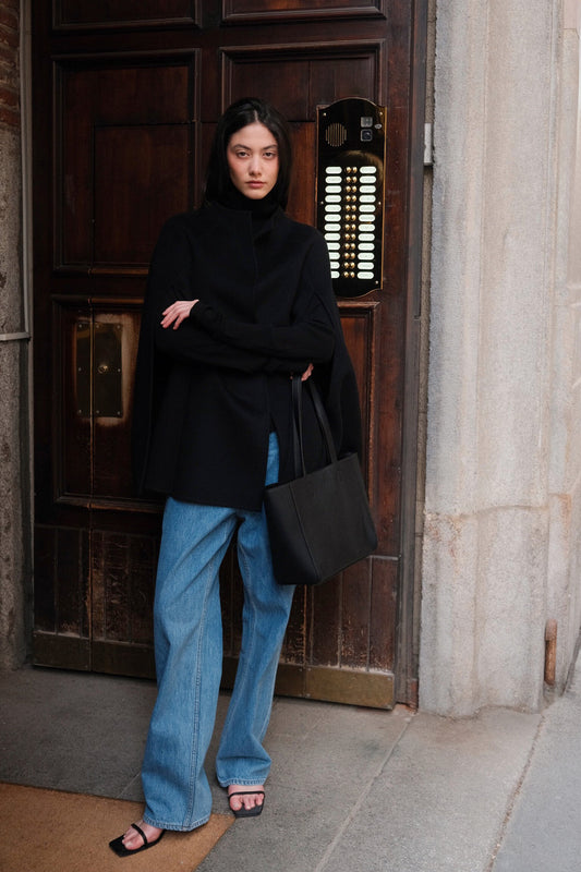 A model stands in front of a dark wooden door wearing a black turtleneck sweater, wide-leg light blue jeans, black sandals, and carrying a large black tote bag.