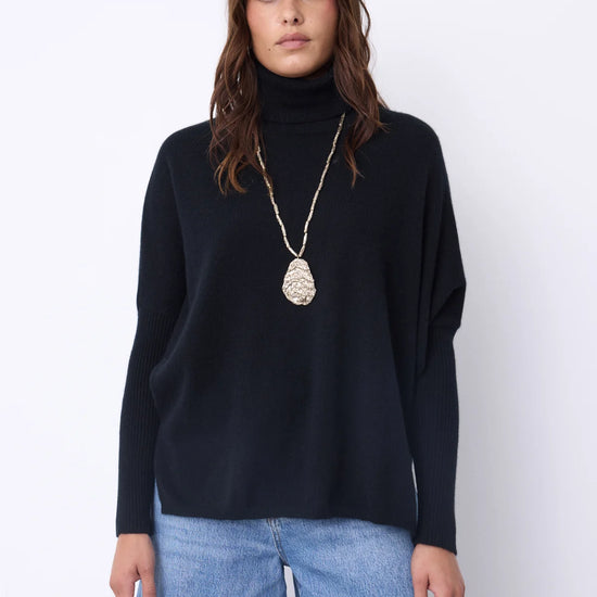 A woman wearing a black long-sleeve turtleneck sweater and light blue jeans, accessorized with a long silver pendant necklace, standing against a plain white studio background.