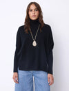 A woman wearing a black long-sleeve turtleneck sweater and light blue jeans, accessorized with a long silver pendant necklace, standing against a plain white studio background.