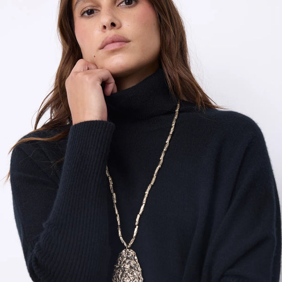 A woman wearing a black ribbed turtleneck sweater and a long silver-toned pendant necklace with a textured teardrop-shaped pendant, posed against a plain white background.