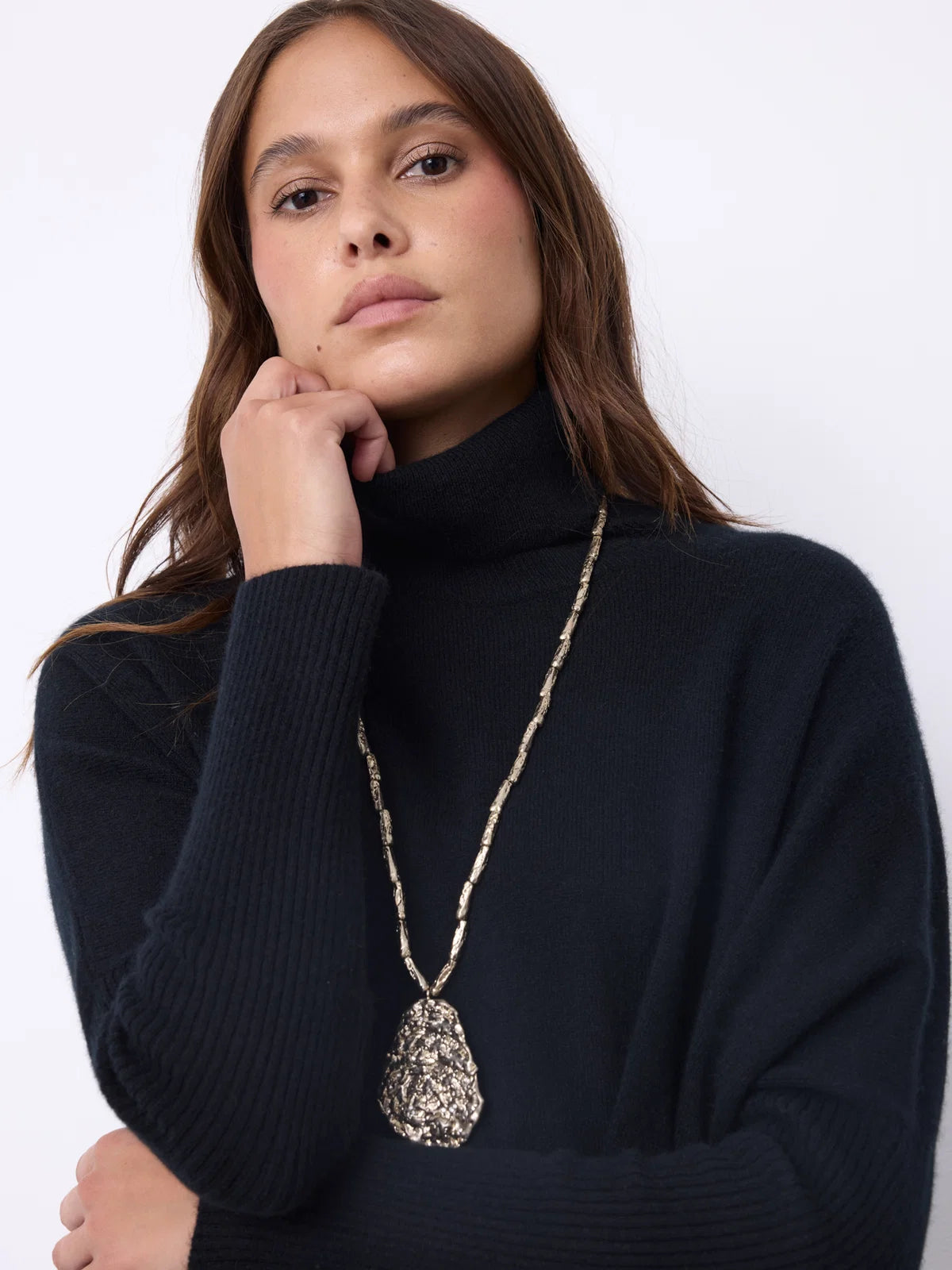 A woman wearing a black ribbed turtleneck sweater and a long silver-toned pendant necklace with a textured teardrop-shaped pendant, posed against a plain white background.