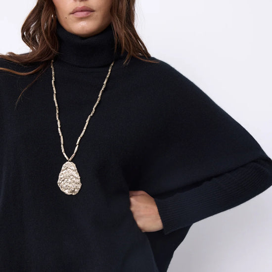 A woman wearing a black turtleneck sweater and a long gold chain necklace with a textured teardrop pendant, photographed against a plain light background.