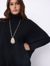 A woman wearing a black turtleneck sweater and a long gold chain necklace with a textured teardrop pendant, photographed against a plain light background.