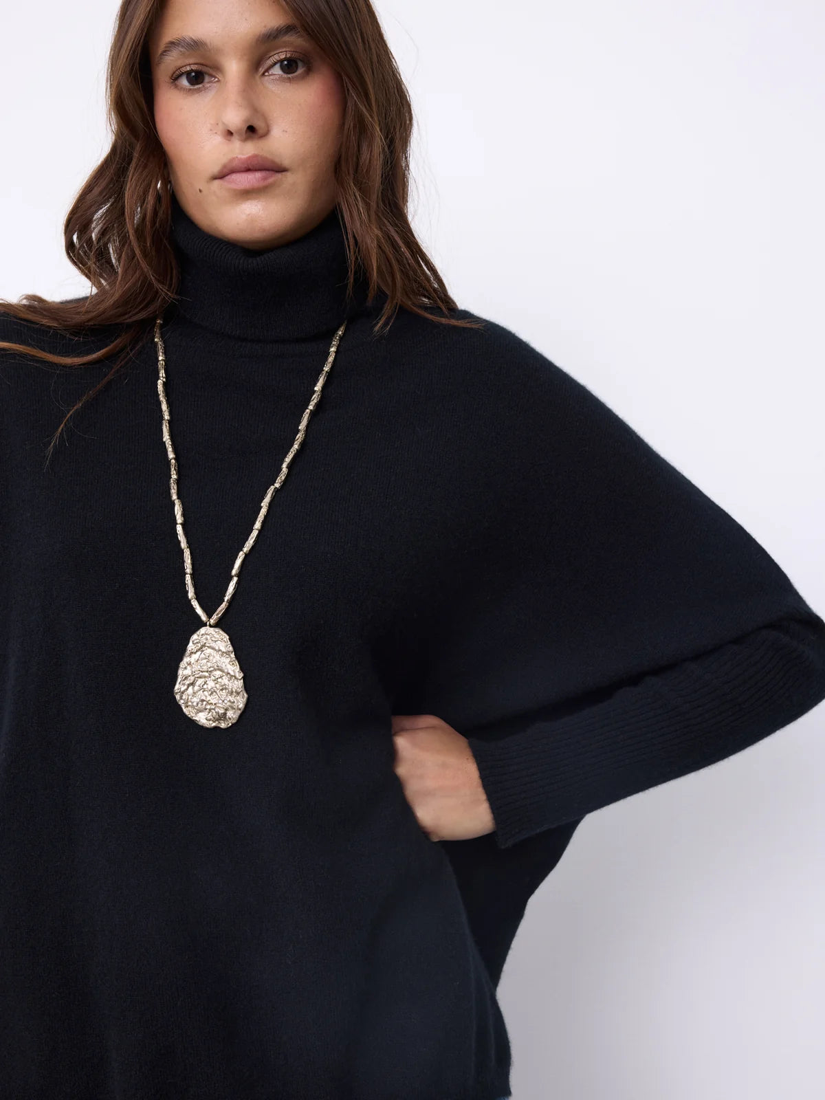A woman wearing a black turtleneck sweater and a long gold chain necklace with a textured teardrop pendant, photographed against a plain light background.