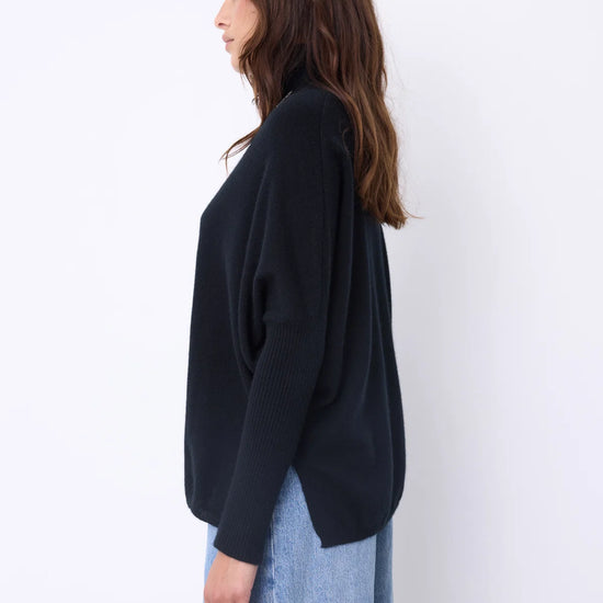 A woman wearing a black long-sleeve turtleneck sweater and light blue jeans, accessorized with a long silver pendant necklace, standing against a plain white studio background.