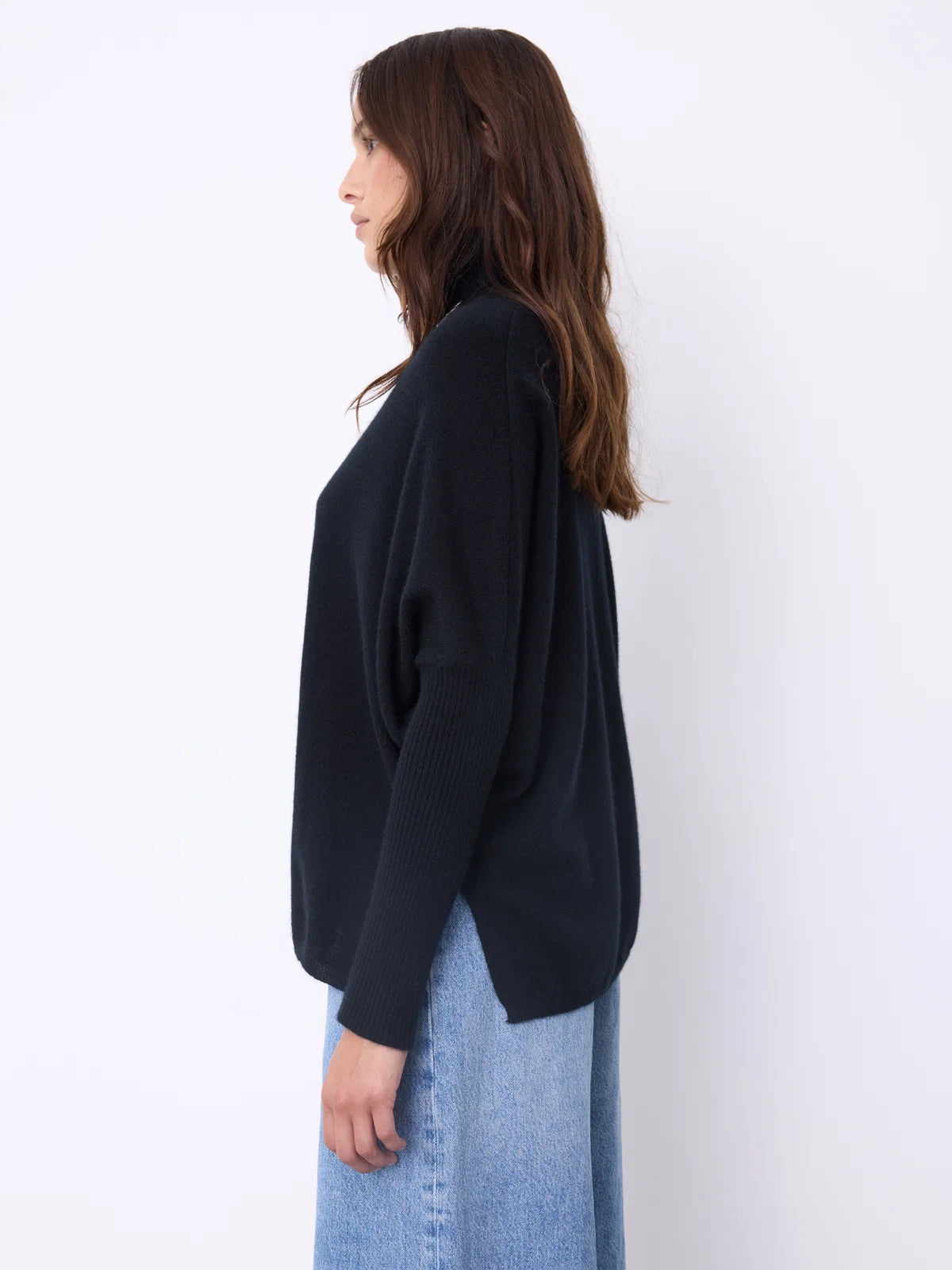 A woman wearing a black long-sleeve turtleneck sweater and light blue jeans, accessorized with a long silver pendant necklace, standing against a plain white studio background.
