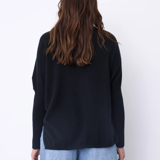 A woman viewed from behind wearing a black long-sleeve knit top and light blue jeans against a plain white background.