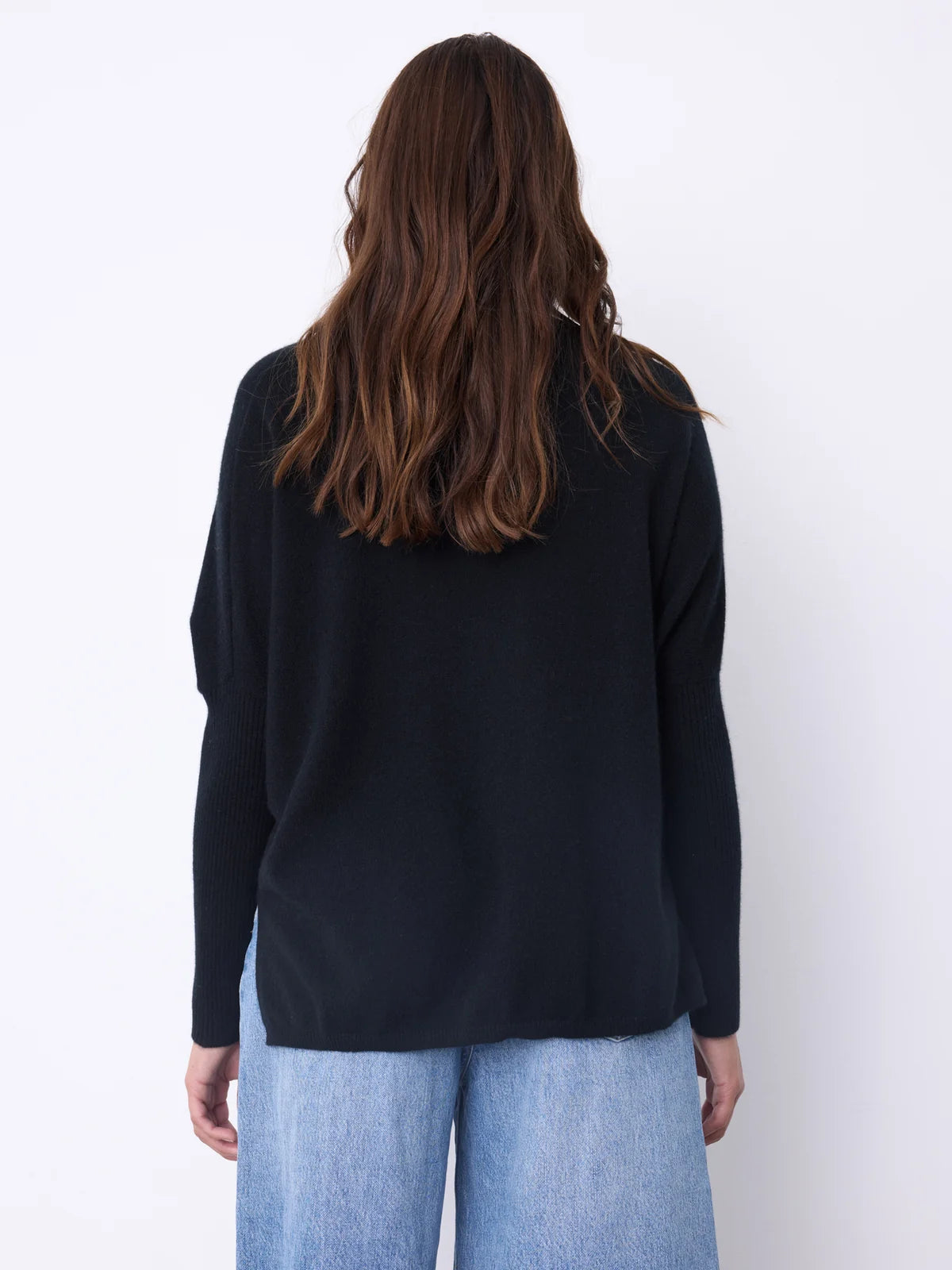 A woman viewed from behind wearing a black long-sleeve knit top and light blue jeans against a plain white background.