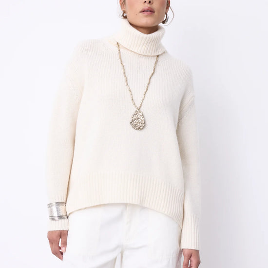 A woman modeling a cream-colored ribbed turtleneck sweater and white trousers against a plain white studio background.
