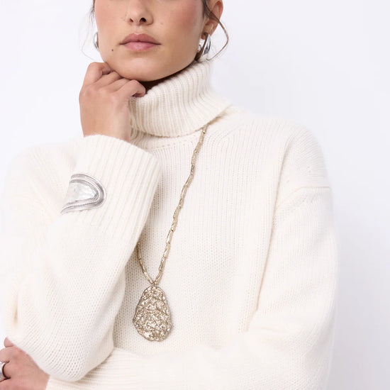 A woman wearing a cream ribbed turtleneck sweater and a long gold chain necklace with a textured teardrop pendant, posed against a clean white background.