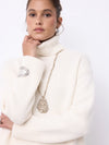A woman wearing a cream ribbed turtleneck sweater and a long gold chain necklace with a textured teardrop pendant, posed against a clean white background.