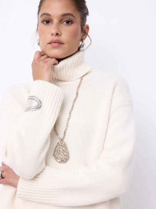 A woman wearing a cream ribbed turtleneck sweater and a long gold chain necklace with a textured teardrop pendant, posed against a clean white background.