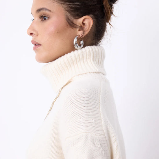 A woman modeling a cream-colored ribbed turtleneck sweater and white trousers against a plain white studio background.