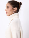 A woman modeling a cream-colored ribbed turtleneck sweater and white trousers against a plain white studio background.