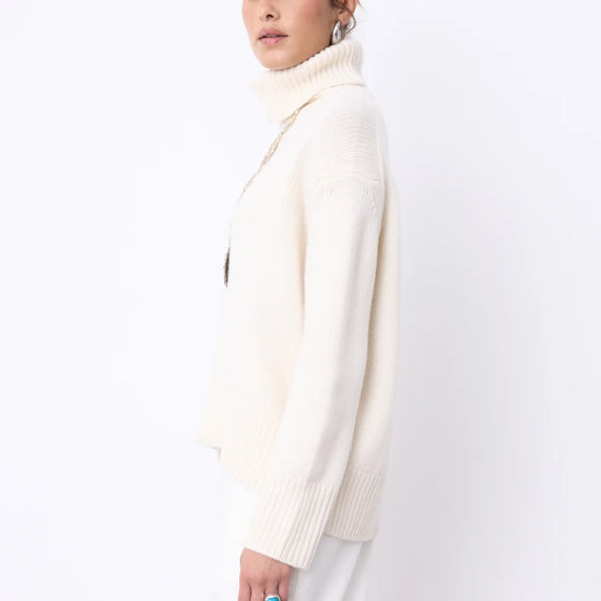 A woman modeling a cream-colored ribbed turtleneck sweater and white trousers against a plain white studio background.