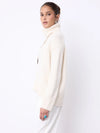 A woman modeling a cream-colored ribbed turtleneck sweater and white trousers against a plain white studio background.