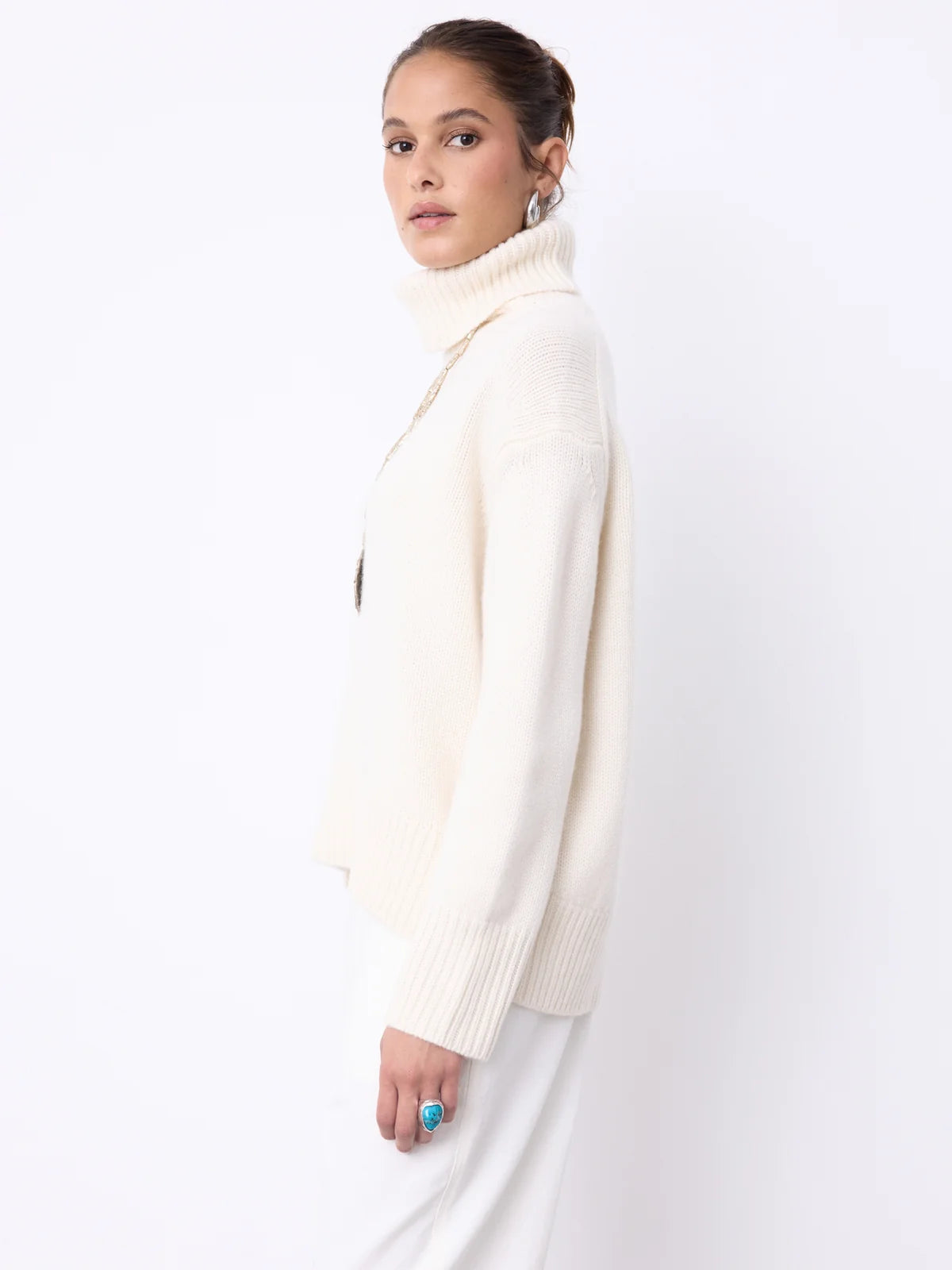 A woman modeling a cream-colored ribbed turtleneck sweater and white trousers against a plain white studio background.