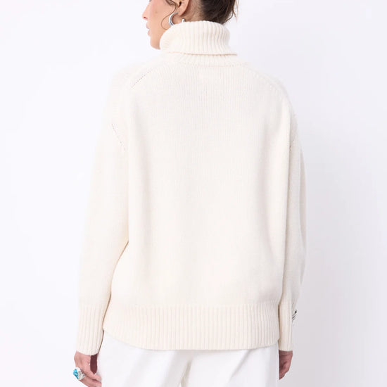 A woman modeling a cream-colored ribbed turtleneck sweater and white trousers against a plain white studio background.