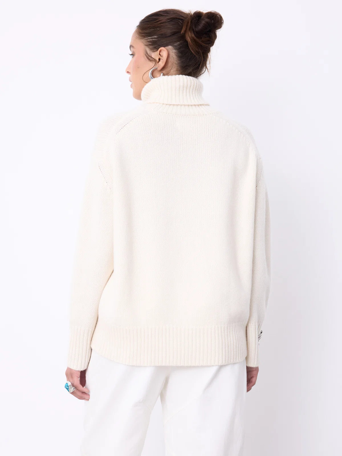 A woman modeling a cream-colored ribbed turtleneck sweater and white trousers against a plain white studio background.