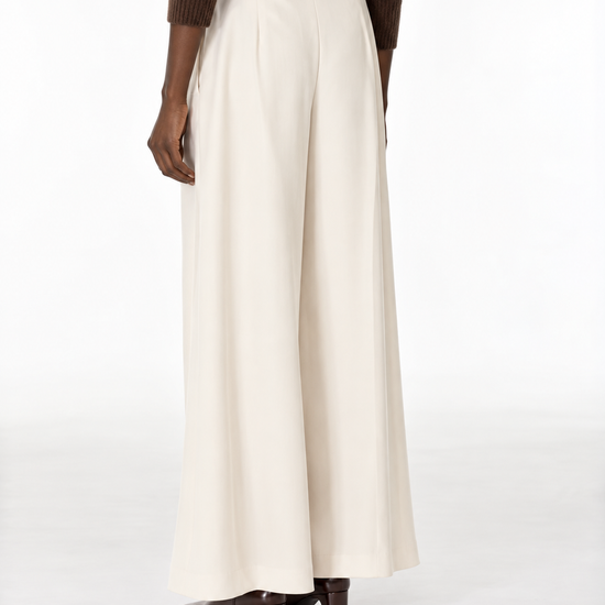 A person shown from the back wearing high-waisted, wide-leg cream trousers, a brown knit sweater, and brown heeled shoes against a plain white studio background.