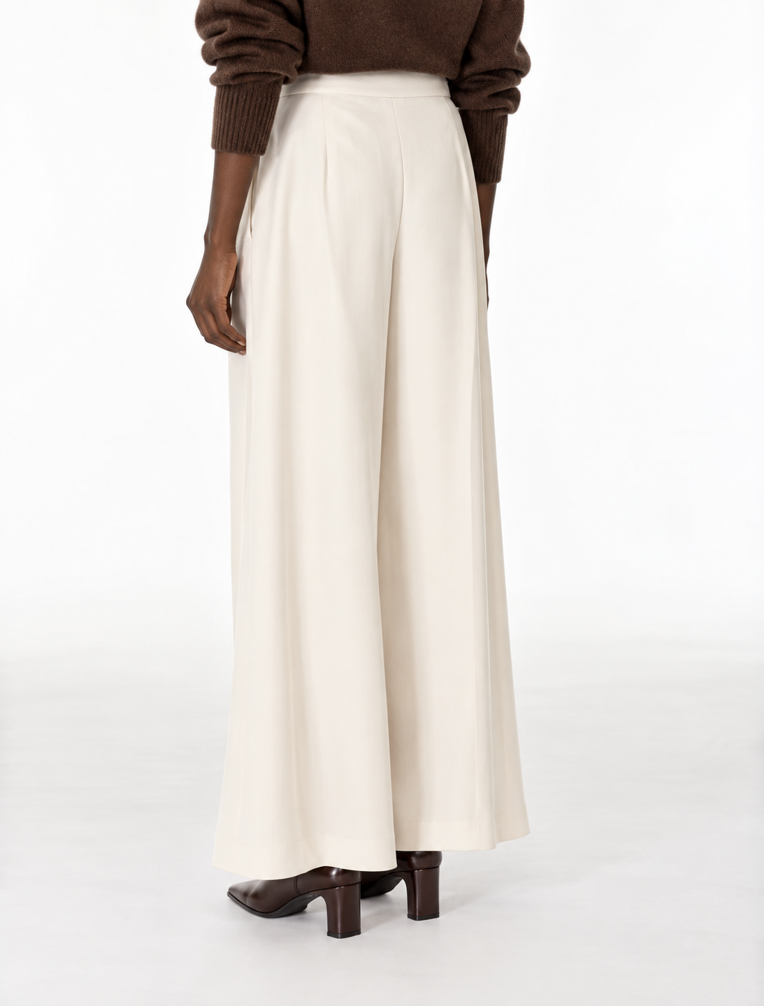 A person shown from the back wearing high-waisted, wide-leg cream trousers, a brown knit sweater, and brown heeled shoes against a plain white studio background.