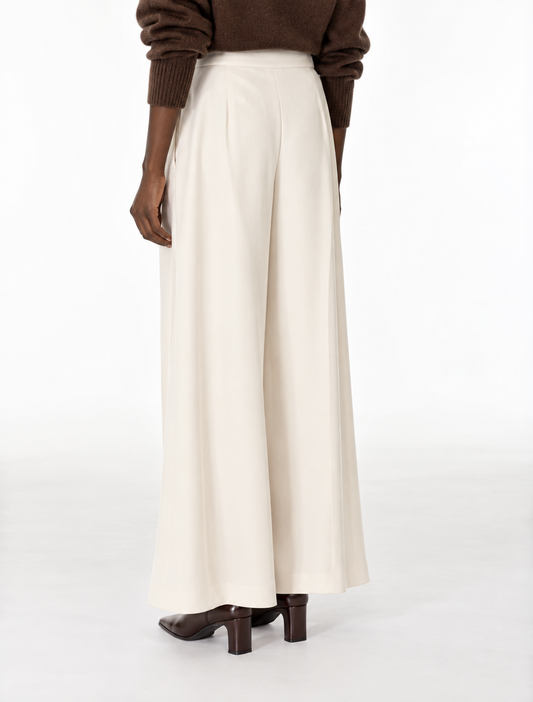 A person shown from the back wearing high-waisted, wide-leg cream trousers, a brown knit sweater, and brown heeled shoes against a plain white studio background.