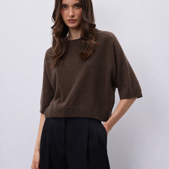 A woman modeling a brown short-sleeve knit top with black wide-leg trousers, standing against a light gray studio background.