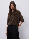 A woman modeling a brown short-sleeve knit top with black wide-leg trousers, standing against a light gray studio background.