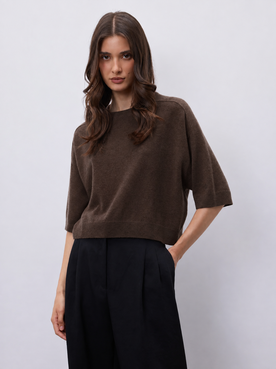A woman modeling a brown short-sleeve knit top with black wide-leg trousers, standing against a light gray studio background.