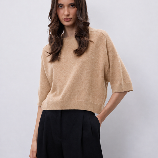 A woman modeling a cropped beige knit sweater with elbow-length sleeves, paired with black wide-leg trousers, against a light gray studio background.
