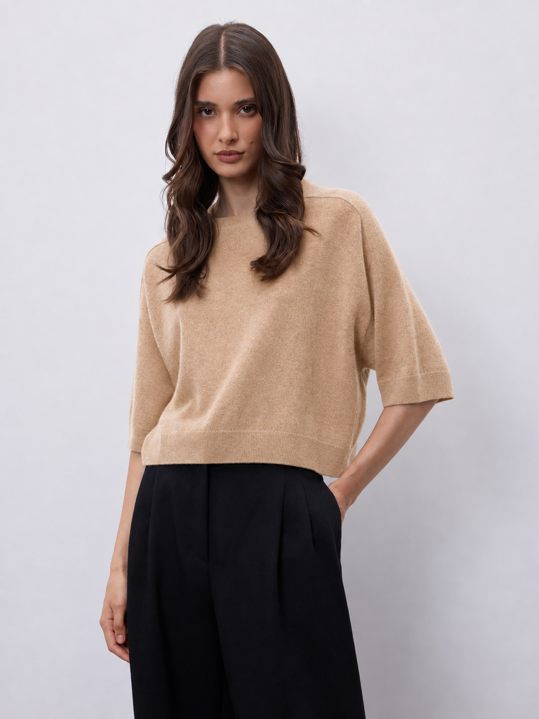 A woman modeling a cropped beige knit sweater with elbow-length sleeves, paired with black wide-leg trousers, against a light gray studio background.