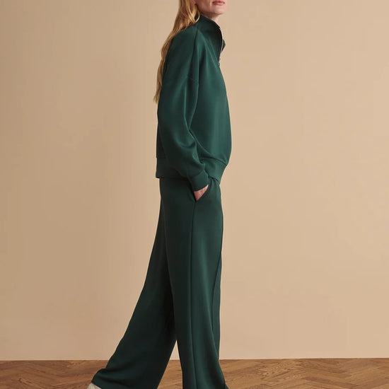 A woman modeling a dark green two-piece lounge set consisting of a long-sleeve mock-neck top and matching wide-leg trousers, paired with white sneakers, set against a warm beige studio background with a wooden floor.